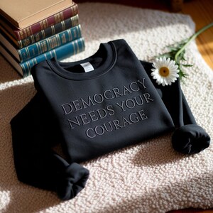 May include: Black sweatshirt with the embroidered text "DEMOCRACY NEEDS YOUR COURAGE." The long-sleeved crewneck is folded on a cream-colored rug, with a stack of books and a daisy nearby.