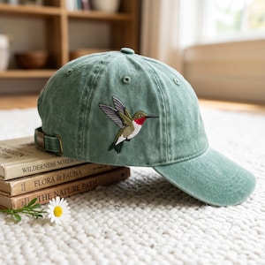 Puede incluir: Una gorra de béisbol color verde salvia con un colibrí bordado en el lateral. El ave tiene alas verdes, pecho blanco y una mancha roja brillante en la garganta. La gorra incluye una correa con hebilla metálica y ojales cosidos para ventilación.