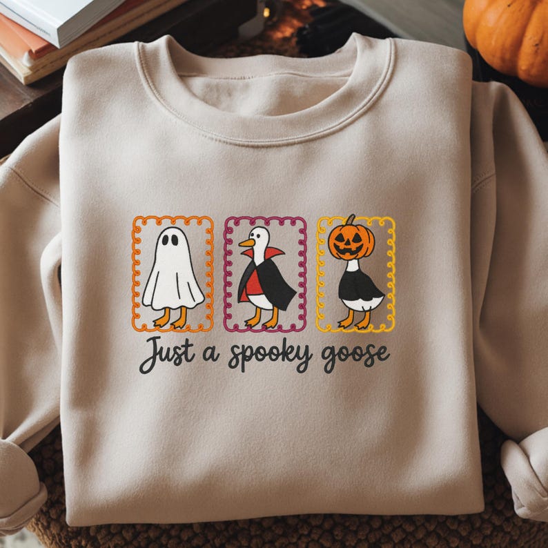 May include: A beige crewneck sweatshirt featuring a graphic of three geese in costumes inside scalloped rectangular frames. The geese are dressed as a ghost, a vampire, and a pumpkin-headed figure. The text "Just a spooky goose" is printed below.