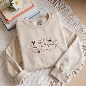 May include: A beige sweatshirt with the phrase "on a side quest" and fantasy-themed graphics in brown. The design includes a dragon, dice, a wand, a wizard hat, and other magical symbols. The sweatshirt is folded on a white rug.