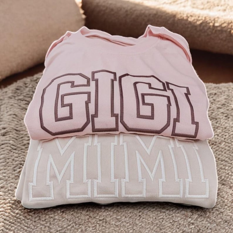 May include: Two pink sweatshirts stacked on top of each other. The top sweatshirt has the word "GIGI" embroidered in brown on the front. The bottom sweatshirt has the word "MIMI" embroidered in white on the front.