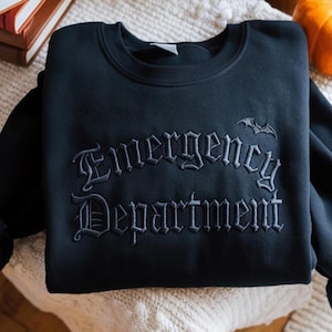 May include: Black sweatshirt with the words "Emergency Department" embroidered in a gothic font. A small bat is embroidered above the word "Emergency". The sweatshirt is folded on a cream-colored surface.