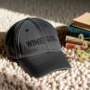 May include: A charcoal grey baseball cap with the embroidered text "WINE GIRL" on the front. The cap is resting on small, white pebbles, with a stack of books and daisies in the background.