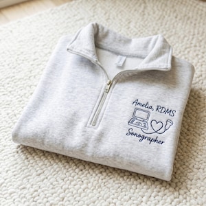 May include: A light gray quarter-zip sweatshirt with a silver zipper. The sweatshirt features the text "Amelia, RDMS Sonographer" along with a sonography machine and heart graphic in navy blue.
