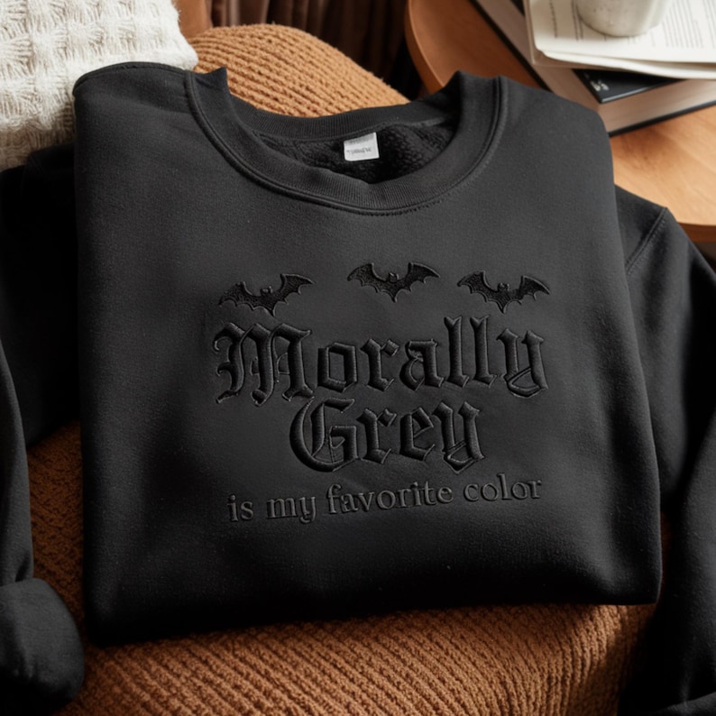 May include: Black sweatshirt with the embroidered phrase "Morally Grey is my favorite color" and three bat silhouettes above the text. The sweatshirt is made of a soft material and is suitable for casual wear.
