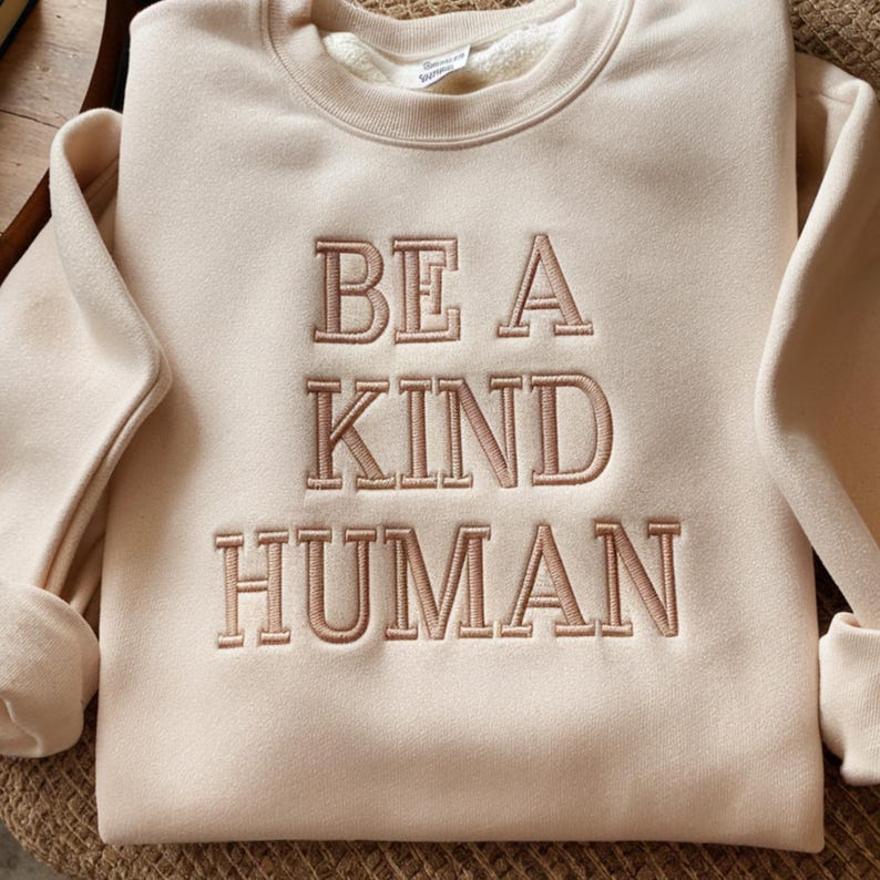 May include: A cream-colored sweatshirt with the phrase "BE A KIND HUMAN" embroidered in brown on the front.