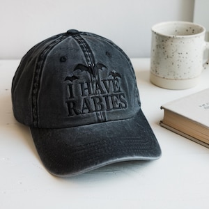 May include: A distressed black baseball cap with the embroidered phrase "I HAVE RABIES" in a gothic font. The cap features a curved brim and a top button. The image also shows a white mug and a book.