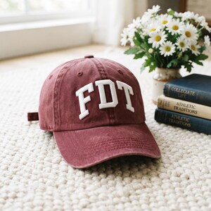 May include: A maroon baseball cap with a distressed finish featuring the white embroidered letters FDT on the front. The cap has a curved brim, a button on the top, and an adjustable strap on the back, showcasing a classic six-panel construction.