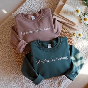 May include: Two crewneck sweatshirts, one dusty rose and one teal, are displayed on a woven basket. The phrase "I'd rather be reading" is embroidered in white on the front of each sweatshirt. A book and daisies are in the background.
