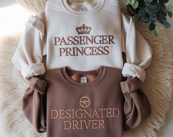May include: Two crewneck sweatshirts, one cream-colored and one brown, are displayed. The cream sweatshirt has "PASSENGER PRINCESS" embroidered in brown with a crown above. The brown sweatshirt says "DESIGNATED DRIVER" with a steering wheel symbol.