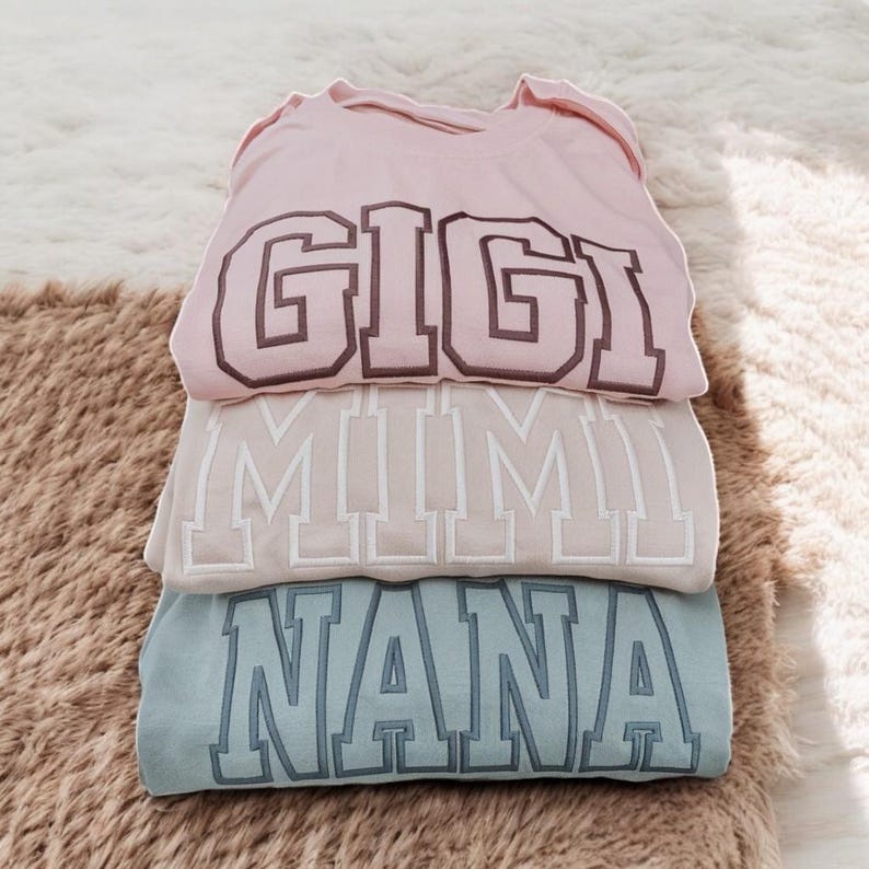 May include: Three sweatshirts stacked on top of each other. The top sweatshirt is pink with the word "GIGI" in brown letters. The middle sweatshirt is beige with the word "MIMI" in white letters. The bottom sweatshirt is light blue with the word "NANA" in dark blue letters.