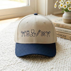 May include: A beige and navy blue baseball cap featuring embroidered medical-themed icons, including a bow, pill bottle, stethoscope, syringe, bandage, and another bow. The cap has a curved brim and a button on top.