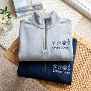 May include: Two quarter-zip sweatshirts, one gray and one navy blue, are stacked on a woven surface. Each sweatshirt features a seashell graphic and the text "Cousins Beach". The gray sweatshirt is on top.