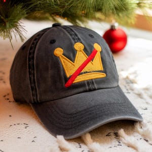 May include: A charcoal gray baseball cap with a distressed finish. The cap features an embroidered gold crown design with a red slash through it. The hat is sitting on a white and cream colored textured surface.