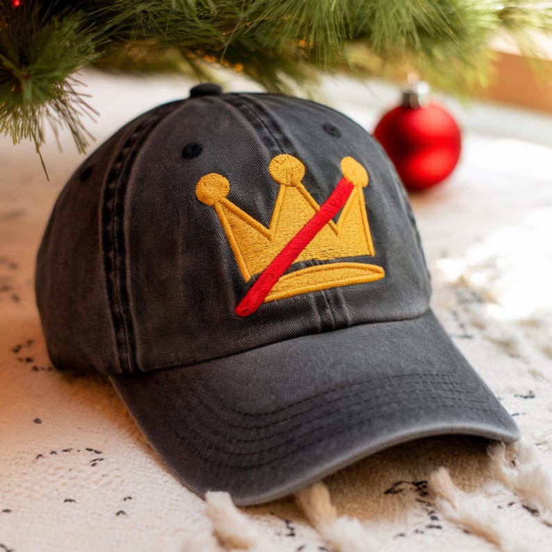May include: A charcoal gray baseball cap with a distressed finish. The cap features an embroidered gold crown design with a red slash through it. The hat is sitting on a white and cream colored textured surface.