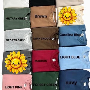 May include: A collection of 12 crew neck sweatshirts in various colors, including white, black, sand, brown, military green, sports grey, dark chocolate, carolina blue, light blue, maroon, forest green, and navy. Some sweatshirts feature a cartoon sunflower graphic.