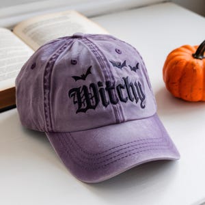 以下が含まれることがあります： 黒い文字で「Witchy」と刺繍された紫色の野球帽。その上に3つの黒いコウモリのシルエットがあります。開いた本と小さなオレンジ色のカボチャが帽子の隣にあります。