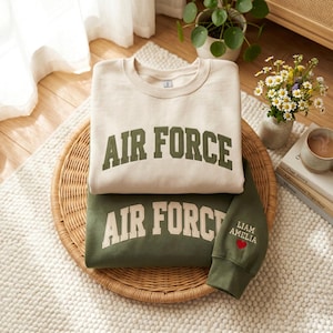 May include: Two sweatshirts, one cream and one olive green, are stacked on a woven tray. The cream sweatshirt has "AIR FORCE" in green block letters. The olive green sweatshirt has the same text and embroidered names and a red heart on the sleeve.