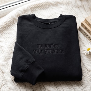 Op de afbeelding: Een zwart sweatshirt met ronde hals en de geborduurde tekst "no gender only stardust" in bijpassend zwart garen. Het kledingstuk heeft lange mouwen met geribbelde boorden en een geribbelde halslijn voor een strak ontwerp.