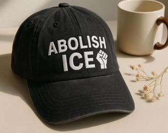 May include: A black baseball cap with the words "ABOLISH ICE" embroidered in white, along with a fist symbol. The cap is made of a soft material and has a curved brim. A beige mug with a brown handle is in the background.
