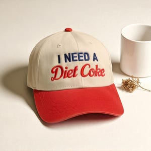 May include: A beige baseball cap with a red brim and top button. The cap features the embroidered phrase "I NEED A Diet Coke" in navy blue and red lettering. A white mug and dried flowers are in the background.