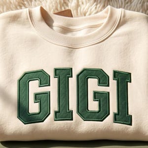 May include: A cream-colored crewneck sweatshirt featuring the word GIGI embroidered in large, dark green block letters across the chest. The garment has a ribbed collar and a smooth, solid-colored fabric construction with a clean, simple design.