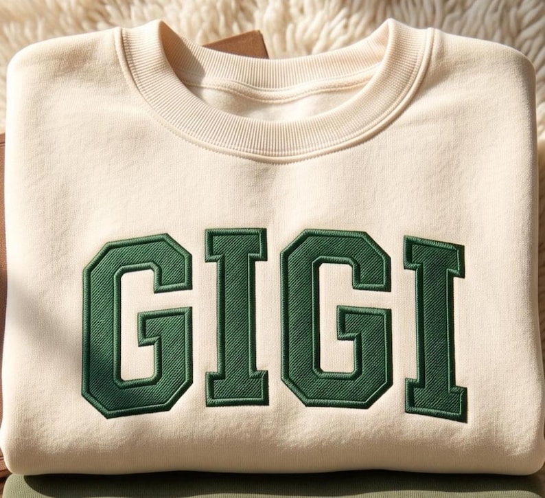 May include: A cream-colored crewneck sweatshirt featuring the word GIGI embroidered in large, dark green block letters across the chest. The garment has a ribbed collar and a smooth, solid-colored fabric construction with a clean, simple design.