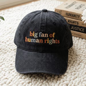 May include: A black baseball cap with a washed-out, vintage texture. The front features embroidered text in orange, yellow, and light blue that reads, "big fan of human rights." The cap has a structured crown, a curved brim, and a button on top.