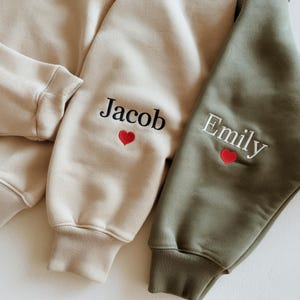 May include: A beige sweatshirt sleeve featuring the embroidered black name Jacob above a small red heart, and an olive green sweatshirt sleeve featuring the embroidered white name Emily above a small red heart, both with ribbed cuffs.