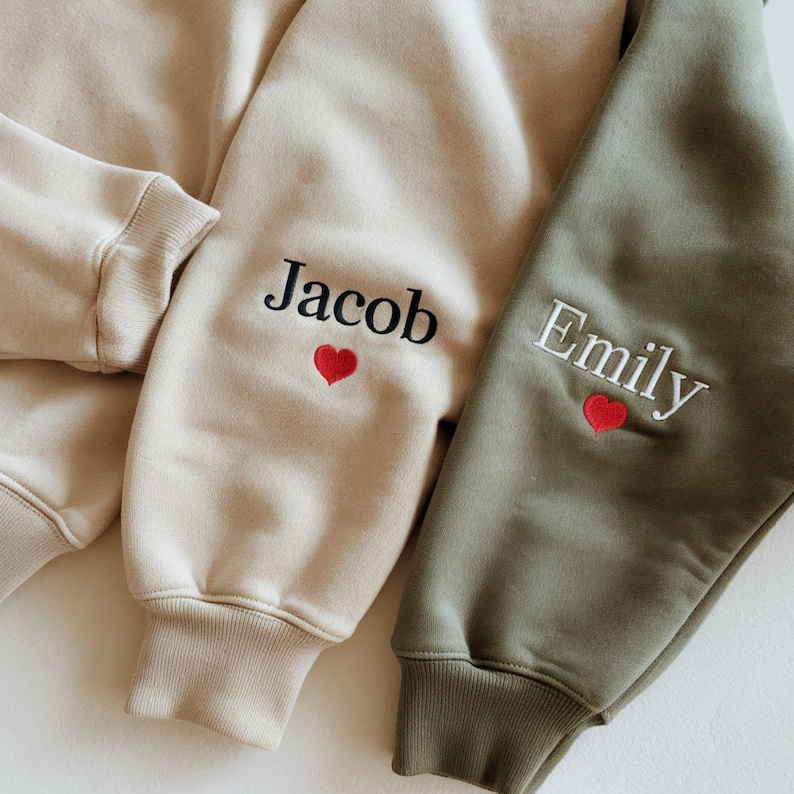 May include: A beige sweatshirt sleeve featuring the embroidered black name Jacob above a small red heart, and an olive green sweatshirt sleeve featuring the embroidered white name Emily above a small red heart, both with ribbed cuffs.