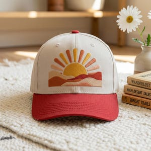 May include: A beige baseball cap with a red brim and button. The cap features an embroidered design of a rising sun over a desert landscape in shades of orange, yellow, and brown.