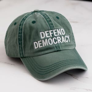 Puede incluir: Una gorra de béisbol verde con las palabras "DEFEND DEMOCRACY" bordadas en blanco en la parte delantera.