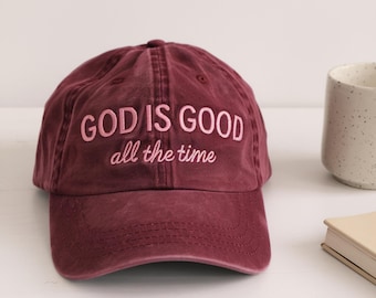 May include: A maroon baseball cap with the embroidered text "GOD IS GOOD all the time" in pink. The cap is made of a soft, worn material. A white mug and a small book are in the background.