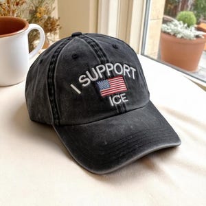 May include: A black baseball cap with the text "I SUPPORT ICE" embroidered in white, along with a small American flag. The cap has a distressed, vintage look.