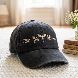 May include: A black baseball cap with a curved brim. The cap features a series of embroidered gold-colored dragons across the front. The hat is sitting on a white rug.