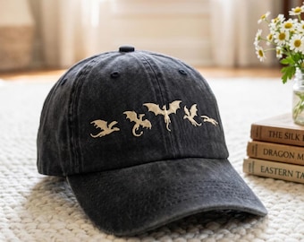 May include: A dark grey baseball cap with a curved peak. The cap has a gold embroidered design of dragons across the front. The hat is on a white rug.
