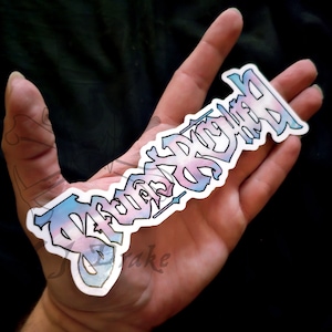 Puede incluir: Una pegatina con la palabra "Transgénero" escrita en una fuente gótica. La pegatina está impresa en un fondo blanco y presenta un esquema de color azul, rosa y blanco.