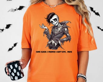 May include: Orange t-shirt with a Halloween-themed graphic. The graphic features a skeleton character in a tattered suit, with the text "COME CLOSER, I PROMISE I DON'T BITE... MUCH." The shirt is paired with a black and white checkered pattern.