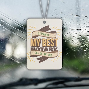 Puede incluir: Un ambientador de coche blanco con el texto "Living My Best Notary Life" en letras marrones y verdes sobre una cinta marrón con estrellas amarillas.