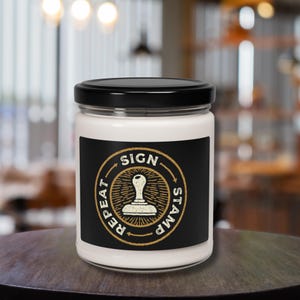 May include: A white candle in a glass jar with a black lid. The candle has a black label with a gold circular design and the words "SIGN", "STAMP", and "REPEAT" around a stamp graphic.