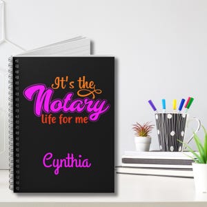以下が含まれることがあります： オレンジとピンクの文字で「It's the Notary life for me」と書かれた黒いスパイラルノート。ノートの下部にピンクで「Cynthia」と書いてあります。