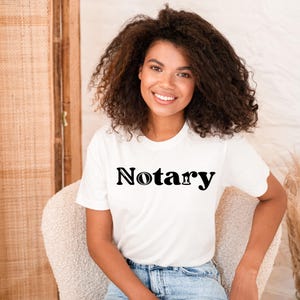 May include: A white t-shirt with the word "Notary" printed in black. The shirt is worn with blue jeans. The person is sitting in a cream-colored chair.