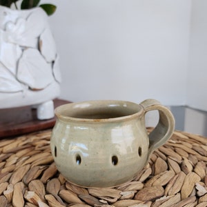 May include: A light green ceramic mug with a handle and small holes around the side. The mug is sitting on a woven wicker surface.