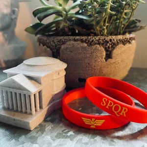 Op de afbeelding: Een miniatuurmodel van een klassiek gebouw met zuilen en een koepeldak, naast twee oranje rubberen armbanden. E&eacute;n armband heeft de letters "SPQR" in goud, en de andere heeft een gouden adembleem. Een vetplant in een pot staat op de achtergrond.