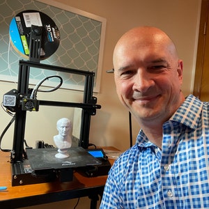 Op de afbeelding: Een zwarte 3D-printer met een witte filamentspoel en een geprinte buste van een persoon. De printer staat op een houten tafel. De persoon op de foto lacht en draagt een blauw-wit geruit overhemd.