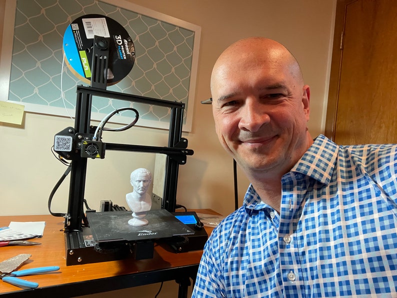 Op de afbeelding: Een zwarte 3D-printer met een witte filamentspoel en een geprinte buste van een persoon. De printer staat op een houten tafel. De persoon op de foto lacht en draagt een blauw-wit geruit overhemd.