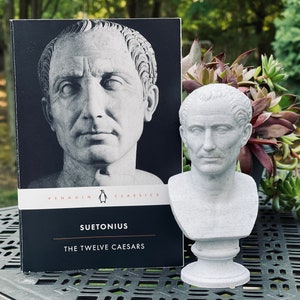 Op de afbeelding: Een grijze buste van een Romeinse keizer staat naast een Penguin Classics-boek met de titel "Suetonius The Twelve Caesars". De boekomslag toont een zwart-wit portret. De buste staat op een voetstuk.