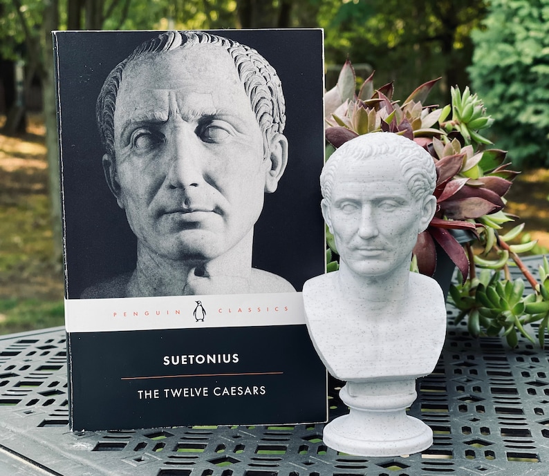 Op de afbeelding: Een grijze buste van een Romeinse keizer staat naast een Penguin Classics-boek met de titel "Suetonius The Twelve Caesars". De boekomslag toont een zwart-wit portret. De buste staat op een voetstuk.