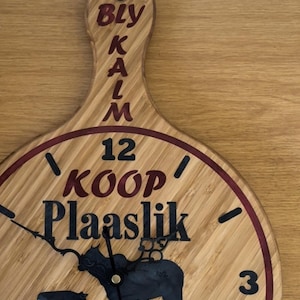 Könnte beinhalten: Eine Holzuhr mit rotem Rand und einem Kuh-Silhouetten-Design. Das Zifferblatt der Uhr zeigt "Koop Plaaslik" und "EST 2023". Der Griff der Uhr trägt die vertikal geschriebenen Worte "Bly Kalm".