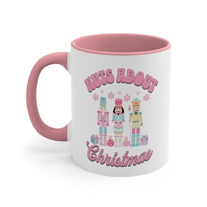Puede incluir: Taza de cerámica blanca con borde y asa de color rosa. La taza presenta un gráfico rosa de tres figuras de cascanueces con el texto "Nuts About Christmas".